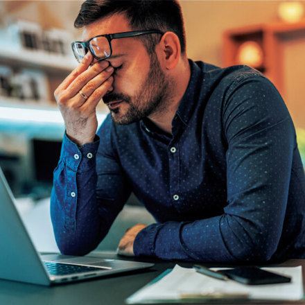 A man sitting at a desk with a laptop, holding his forehead in apparent frustration. He wears a blue shirt and glasses, and a cup and papers are on the desk. The background has warm lighting and blurred shelves.