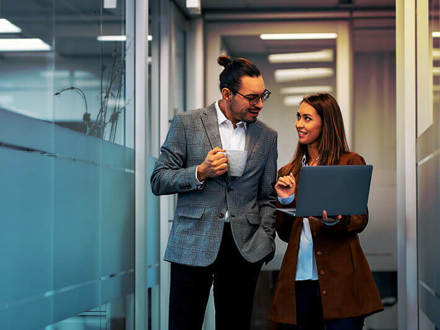 A man holding a coffee mug and a woman holding a laptop stand in a modern office hallway, engaged in conversation. They both wear business attire. The hallway is bright, with glass walls and overhead lights.