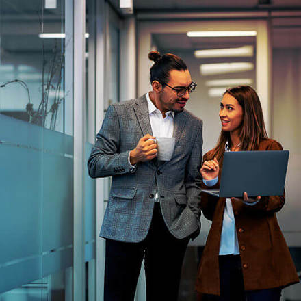 A man holding a coffee mug and a woman holding a laptop stand in a modern office hallway, engaged in conversation. They both wear business attire. The hallway is bright, with glass walls and overhead lights.