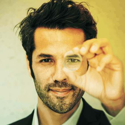 Handsome man holding a lens up to his left eye and looking through it.