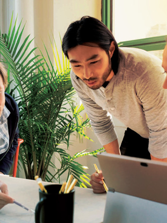 Three people are working together around a table in a bright room with green window frames and plants. One person points at a document, while another looks on attentively, and the third person engages with a laptop.