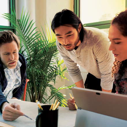 Three people are working together around a table in a bright room with green window frames and plants. One person points at a document, while another looks on attentively, and the third person engages with a laptop.