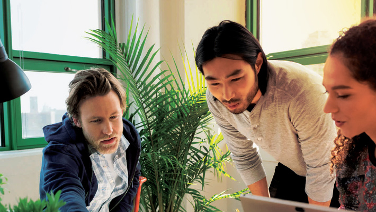 Three people are working together around a table in a bright room with green window frames and plants. One person points at a document, while another looks on attentively, and the third person engages with a laptop.