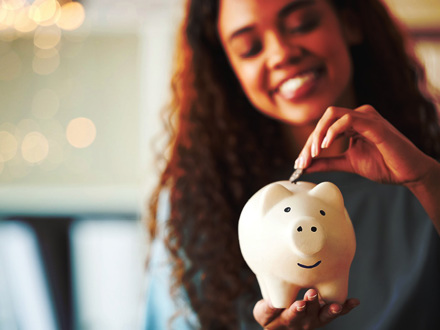 A person with long curly hair smiles while placing a coin into a white piggy bank. The background is softly blurred with warm lighting, emphasizing the piggy bank in focus.