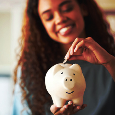 A person with long curly hair smiles while placing a coin into a white piggy bank. The background is softly blurred with warm lighting, emphasizing the piggy bank in focus.