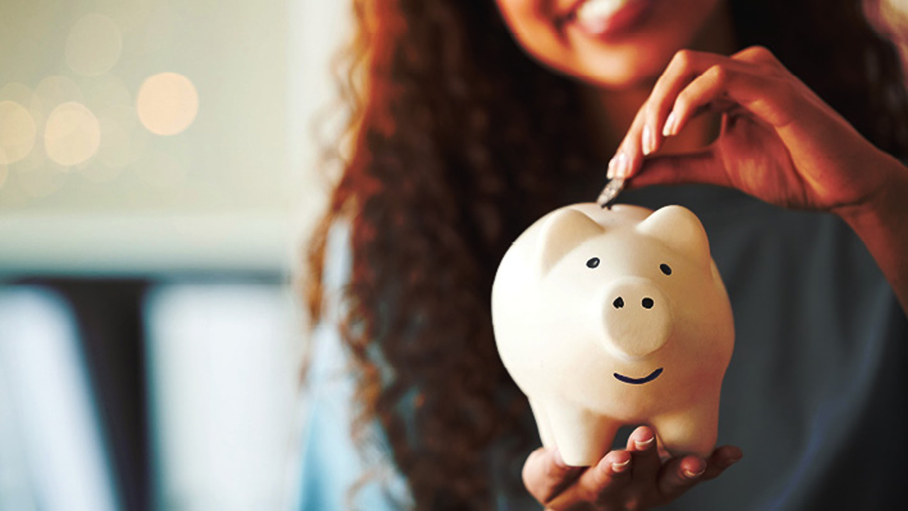 A person with long curly hair smiles while placing a coin into a white piggy bank. The background is softly blurred with warm lighting, emphasizing the piggy bank in focus.