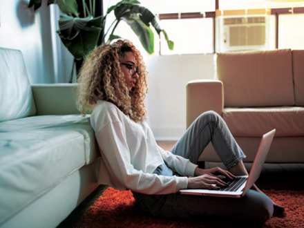 A person with curly hair sits on the floor, leaning against a couch, and types on a laptop. They are in a brightly lit room with a large window and a plant in the background.