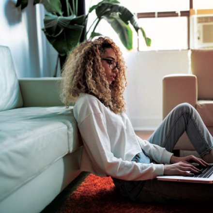 A person with curly hair sits on the floor, leaning against a couch, and types on a laptop. They are in a brightly lit room with a large window and a plant in the background.