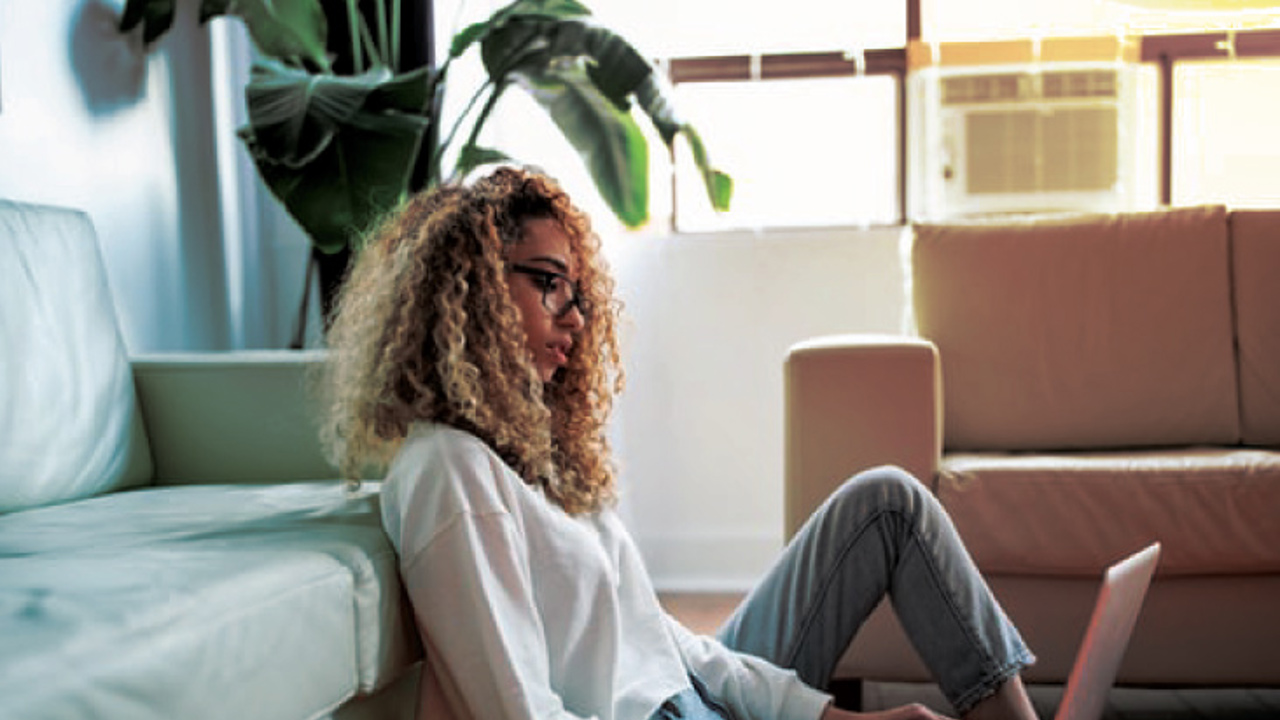 A person with curly hair sits on the floor, leaning against a couch, and types on a laptop. They are in a brightly lit room with a large window and a plant in the background.