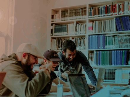 Four people are gathered around a table, engaged in a lively discussion while looking at a laptop. They are surrounded by bookshelves filled with binders and books. Natural light filters in from a nearby window, creating a warm atmosphere.