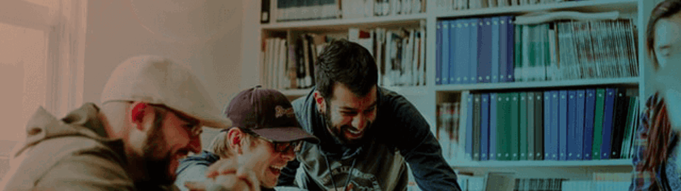 Four people are gathered around a table, engaged in a lively discussion while looking at a laptop. They are surrounded by bookshelves filled with binders and books. Natural light filters in from a nearby window, creating a warm atmosphere.