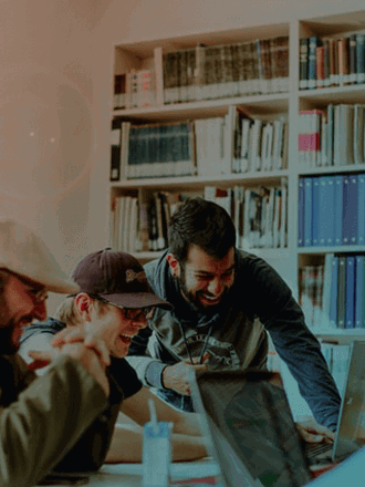 Four people are gathered around a table, engaged in a lively discussion while looking at a laptop. They are surrounded by bookshelves filled with binders and books. Natural light filters in from a nearby window, creating a warm atmosphere.