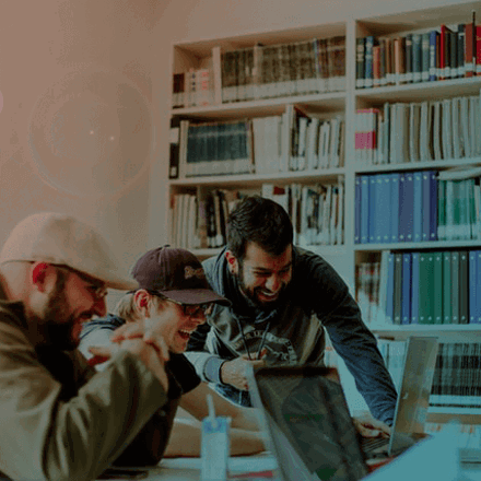 Four people are gathered around a table, engaged in a lively discussion while looking at a laptop. They are surrounded by bookshelves filled with binders and books. Natural light filters in from a nearby window, creating a warm atmosphere.