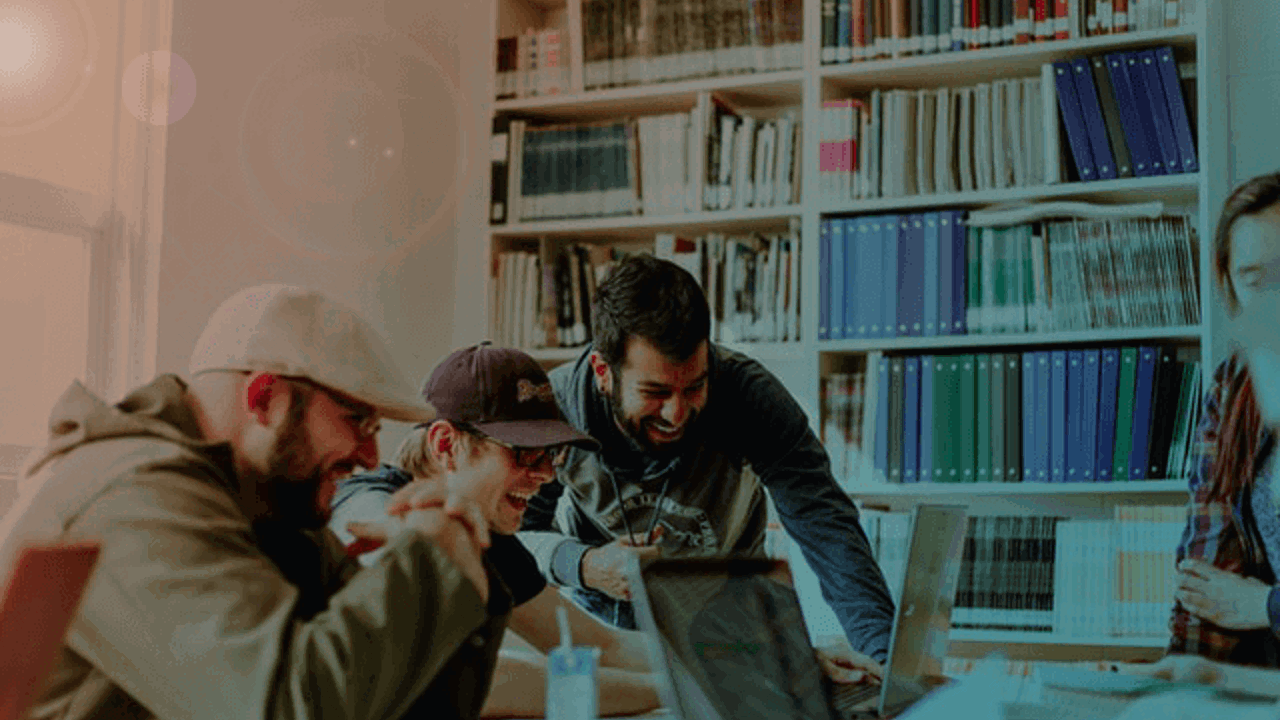 Four people are gathered around a table, engaged in a lively discussion while looking at a laptop. They are surrounded by bookshelves filled with binders and books. Natural light filters in from a nearby window, creating a warm atmosphere.