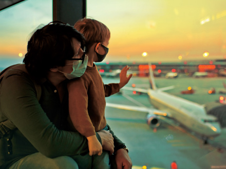 A man and a child wearing masks sit at an airport window, watching planes on the tarmac during sunset. The child waves at an airplane, while the sky glows orange and pink.