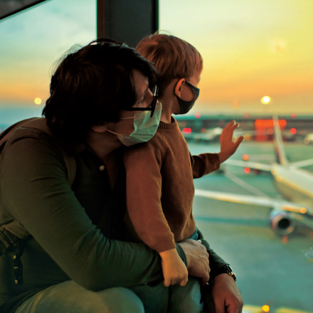 A man and a child wearing masks sit at an airport window, watching planes on the tarmac during sunset. The child waves at an airplane, while the sky glows orange and pink.