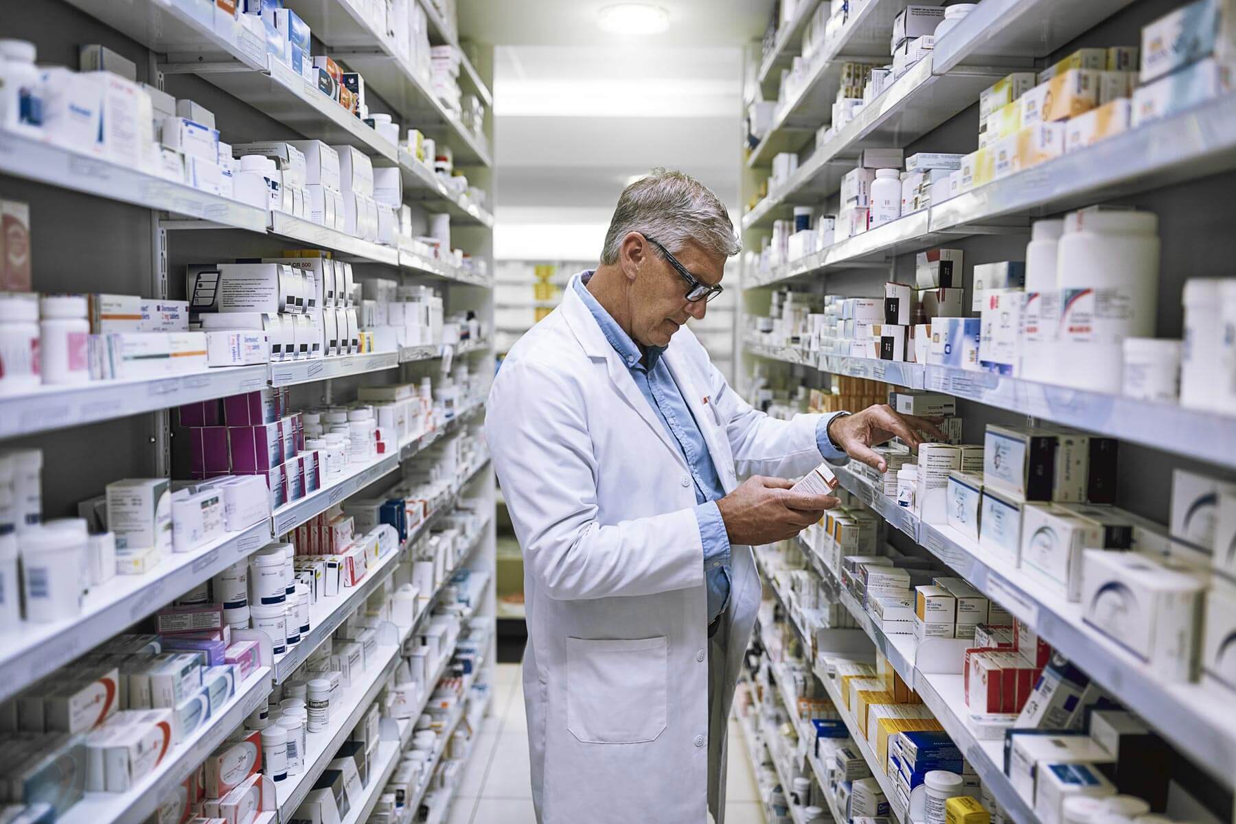 A pharmacist in a white coat and glasses examines medication in a well-stocked pharmacy aisle. Shelves are lined with various boxes and bottles of pharmaceutical products.