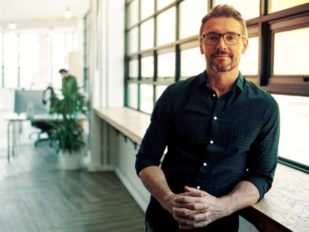 A man with glasses and a beard stands confidently, leaning against a windowsill in a modern, sunlit office. He is wearing a dark, patterned shirt, and the office features large windows and a person working at a desk in the background.