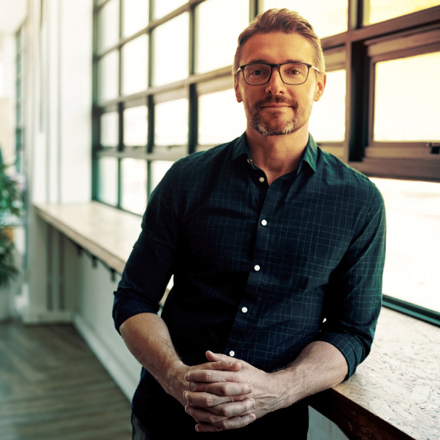 A man with glasses and a beard stands confidently, leaning against a windowsill in a modern, sunlit office. He is wearing a dark, patterned shirt, and the office features large windows and a person working at a desk in the background.