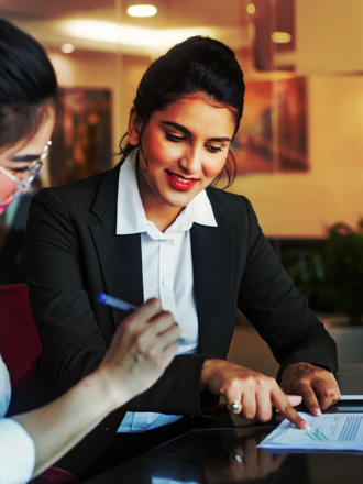 Two women are sitting at a table in an office setting. One is reviewing a document while the other points to a laptop screen. They are both dressed in business attire and appear to be engaged in a discussion.