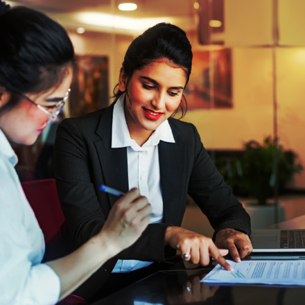 Two women are sitting at a table in an office setting. One is reviewing a document while the other points to a laptop screen. They are both dressed in business attire and appear to be engaged in a discussion.