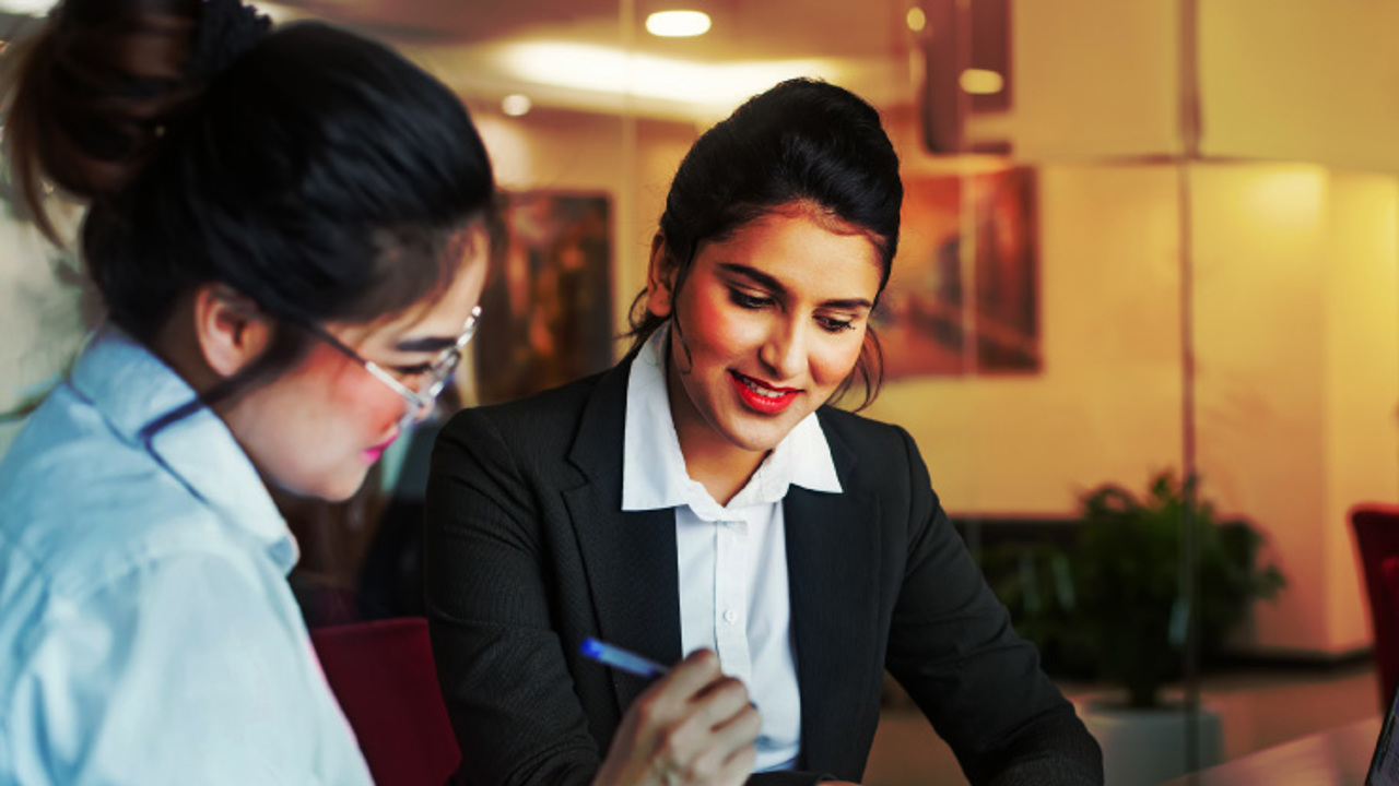 Two women are sitting at a table in an office setting. One is reviewing a document while the other points to a laptop screen. They are both dressed in business attire and appear to be engaged in a discussion.