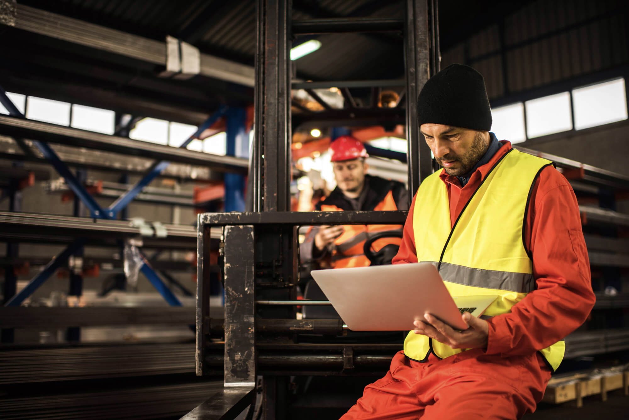 A worker in a yellow safety vest and beanie uses a laptop in a warehouse. In the background, another worker in red protective gear operates machinery surrounded by metal beams.