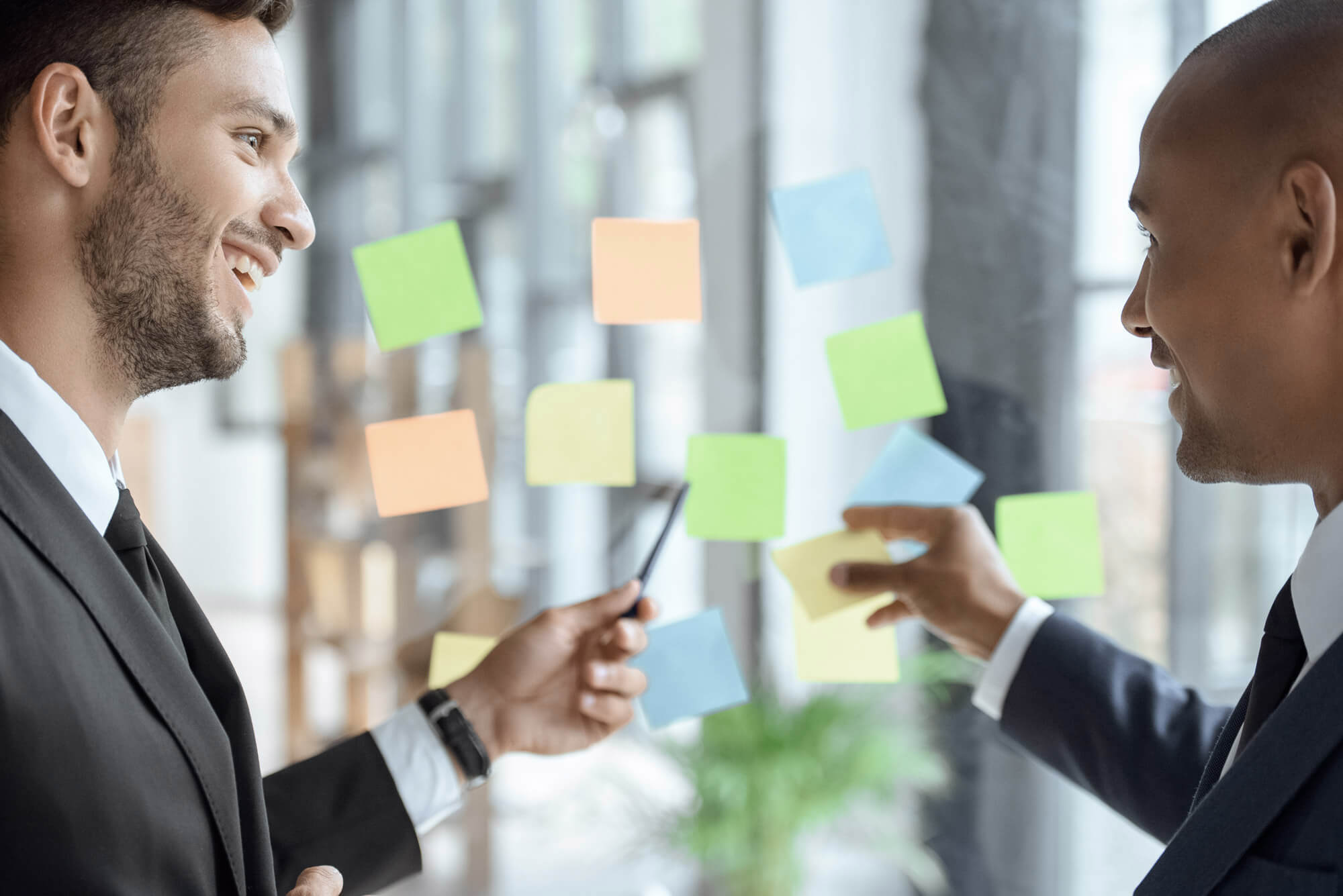 Two men in suits are discussing ideas in an office setting. They are standing in front of a glass wall covered with colorful sticky notes, smiling and gesturing as they collaborate.