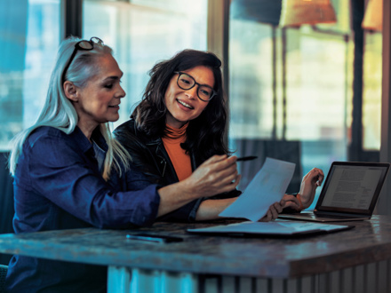 Two women sit at a table in an office setting, engaged in a discussion. One is holding a document and pen, while the other, with a laptop open in front of her, listens attentively. Both appear focused and collaborative.