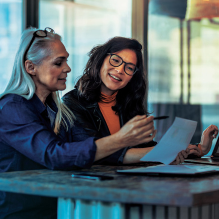 Two women sit at a table in an office setting, engaged in a discussion. One is holding a document and pen, while the other, with a laptop open in front of her, listens attentively. Both appear focused and collaborative.