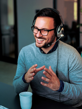 A smiling man with glasses and a headset sits at a desk, gesturing with his hands while looking at a computer screen. A cup is in front of him, and blurred office furniture is in the background.