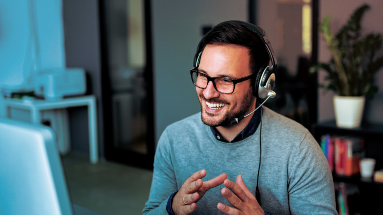 A smiling man with glasses and a headset sits at a desk, gesturing with his hands while looking at a computer screen. A cup is in front of him, and blurred office furniture is in the background.