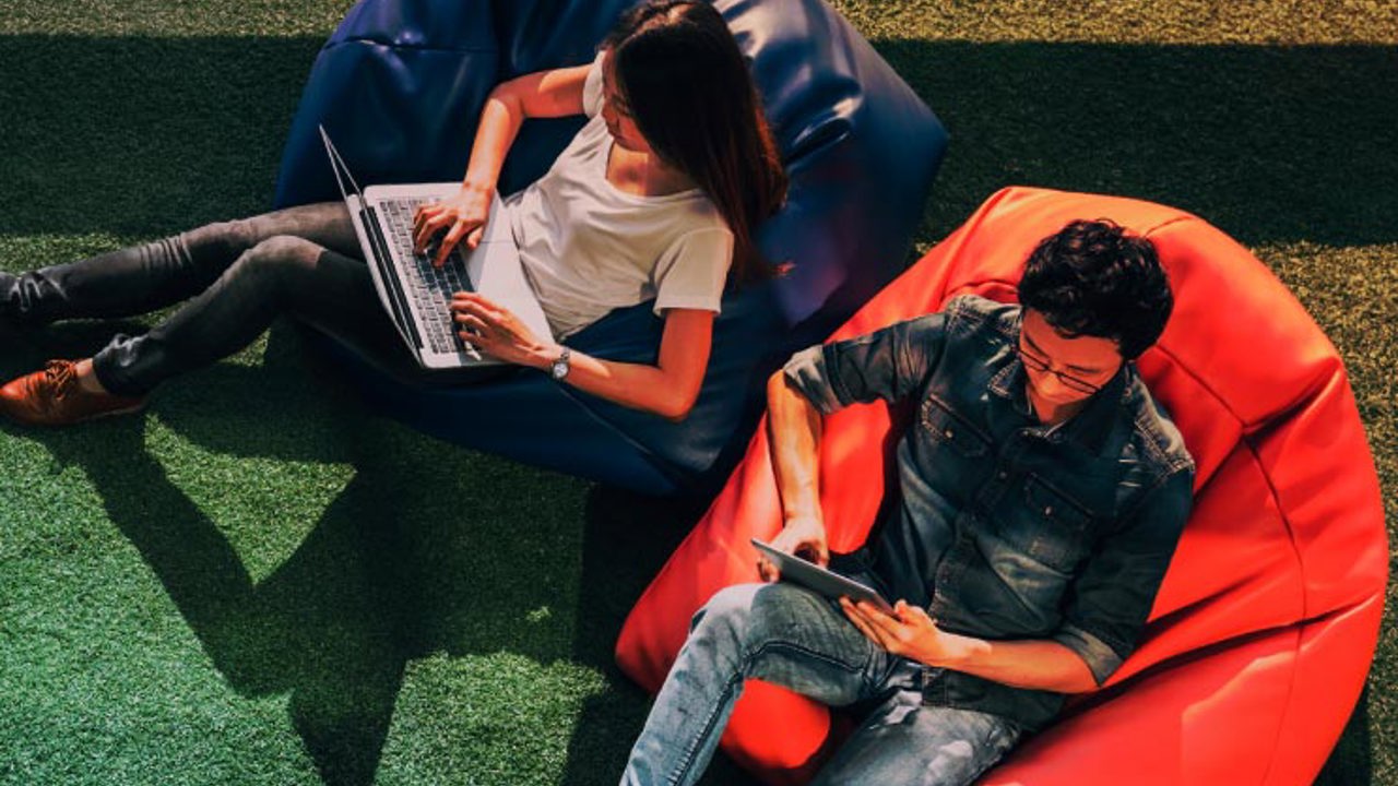 Young Asian couple using laptop notebook and digital tablet together in modern public park, sit on bean bag, top view with copy space on grass.