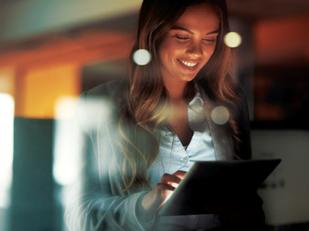 A woman smiles while using a tablet. She is indoors, and the foreground shows reflections of circular lights, creating a warm and focused atmosphere.