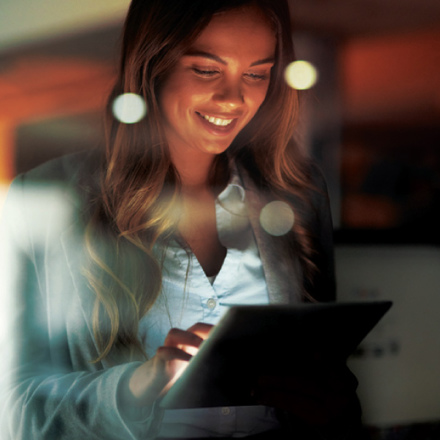 A woman smiles while using a tablet. She is indoors, and the foreground shows reflections of circular lights, creating a warm and focused atmosphere.