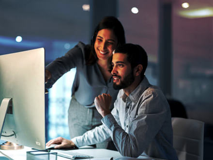 A woman and a man are working together at a computer. The woman is smiling and pointing at the screen, while the man is seated, focusing on the monitor. The atmosphere is collaborative and positive.