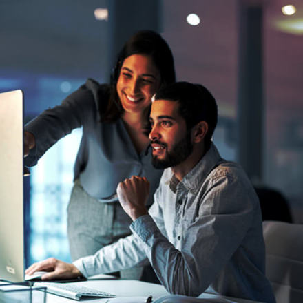 A woman and a man are working together at a computer. The woman is smiling and pointing at the screen, while the man is seated, focusing on the monitor. The atmosphere is collaborative and positive.