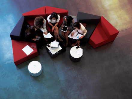 A group of five people sitting on a curved sofa in a modern office setting, discussing with tablets and papers. The furniture is black and red, and there are small round tables nearby. The view is from above, showing a polished concrete floor.