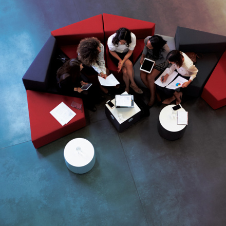 A group of five people sitting on a curved sofa in a modern office setting, discussing with tablets and papers. The furniture is black and red, and there are small round tables nearby. The view is from above, showing a polished concrete floor.