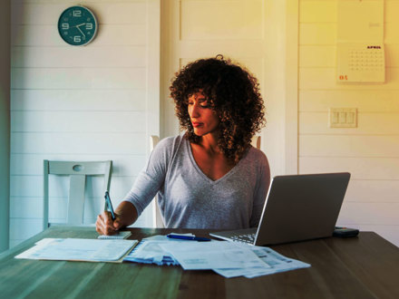 A woman sits at her dining room table with laptop and financial reports doing her monthly budget. She is writing down budgets as she works on her computer to do monthly finances, pay taxes and save money for the future.