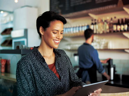 A person with short hair smiles while holding a tablet in a cozy coffee shop. Shelves with bottles and a barista in the background create a warm atmosphere. The person is wearing a polka dot shirt.