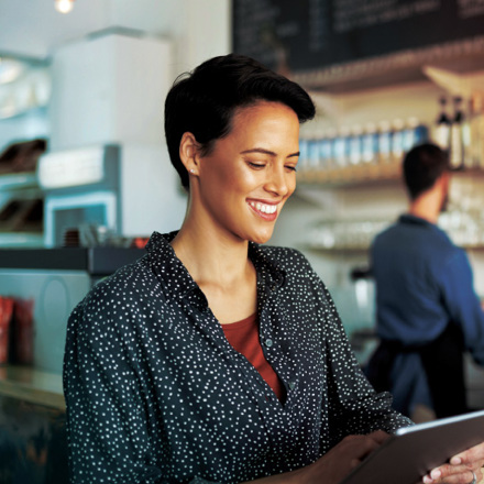 A person with short hair smiles while holding a tablet in a cozy coffee shop. Shelves with bottles and a barista in the background create a warm atmosphere. The person is wearing a polka dot shirt.