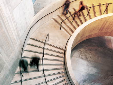 Blurred motion of people ascending and descending a spiral staircase with concrete walls. The image captures the dynamic flow of movement, highlighted by the curvature of the architecture and the soft lighting.