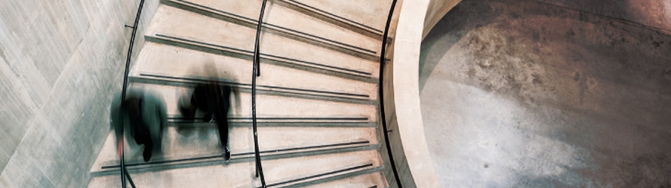 Blurred motion of people ascending and descending a spiral staircase with concrete walls. The image captures the dynamic flow of movement, highlighted by the curvature of the architecture and the soft lighting.