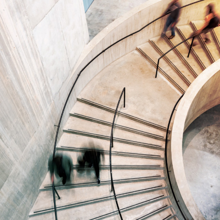 Blurred motion of people ascending and descending a spiral staircase with concrete walls. The image captures the dynamic flow of movement, highlighted by the curvature of the architecture and the soft lighting.