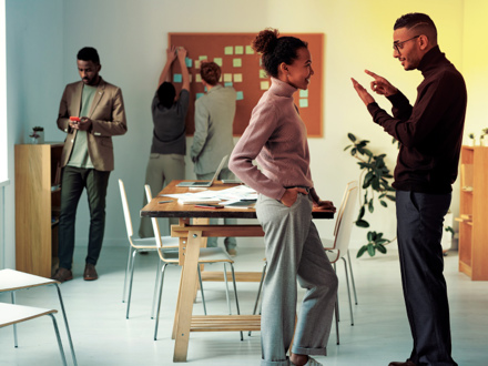 A group of people in an office setting. Two individuals are having a discussion in the foreground while another is looking at a phone. In the background, two people are placing sticky notes on a wall-mounted board. Chairs and tables are around.