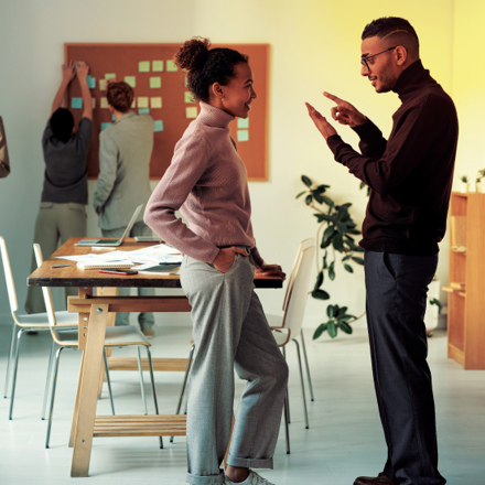 A group of people in an office setting. Two individuals are having a discussion in the foreground while another is looking at a phone. In the background, two people are placing sticky notes on a wall-mounted board. Chairs and tables are around.