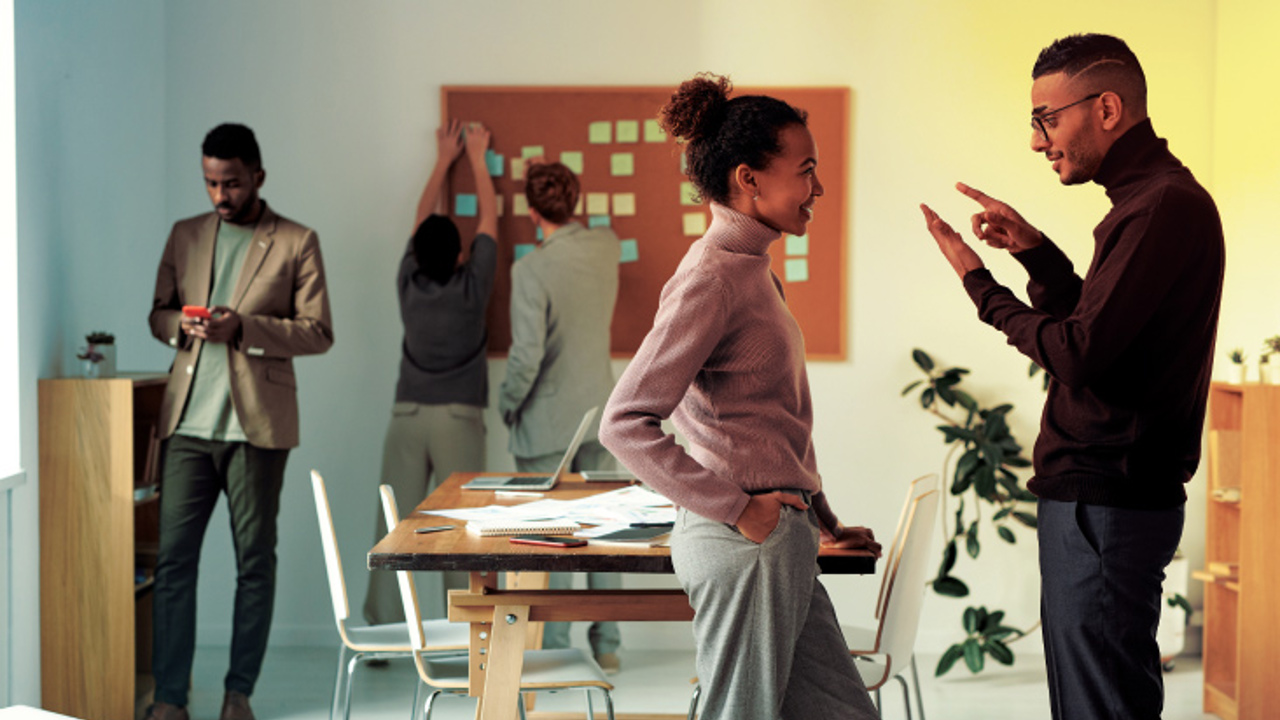 A group of people in an office setting. Two individuals are having a discussion in the foreground while another is looking at a phone. In the background, two people are placing sticky notes on a wall-mounted board. Chairs and tables are around.