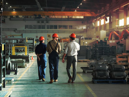 Three workers in helmets and overalls walk through a spacious, well-lit factory. Metal parts and equipment are organized on pallets and shelves. A large overhead crane is visible above, and a safety cone is placed near the path.