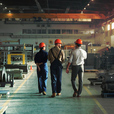 Three workers in helmets and overalls walk through a spacious, well-lit factory. Metal parts and equipment are organized on pallets and shelves. A large overhead crane is visible above, and a safety cone is placed near the path.